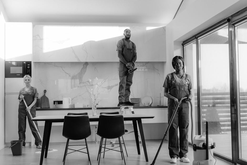 Three professional cleaners from Cleaners Cleaning NW5 are performing surface cleaning and deep cleaning tasks in a modern kitchen with a marble backsplash. One cleaner stands on a step ladder carefully scrubbing high on the wall, while another uses a mop on the wooden floor near a dining table with black chairs. The third cleaner is sweeping the floor with a vacuum cleaner. The kitchen features large sliding glass doors that allow natural light to illuminate the spotless, shiny surfaces, including the polished wooden floors, sleek marble backsplash, and stainless steel appliances. The scene emphasizes thorough cleaning and sanitisation of domestic living spaces, showcasing the company's expertise in maintaining hygiene and cleanliness in residential environments.