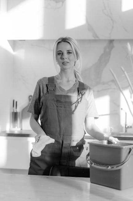 A young woman with light hair tied back, wearing a casual T-shirt and overalls, stands in a modern kitchen with a clean, marble-patterned wall behind her. She is holding a cleaning cloth in one hand and appears to be preparing for surface cleaning or sanitisation. In front of her on the countertop is a container, possibly filled with cleaning supplies or water, with a mop handle visible in the background. The kitchen floor is smooth and tidy, and natural light illuminates the space, emphasizing the cleanliness and hygiene of the area. The scene depicts domestic cleaning processes, highlighting a professional approach by Cleaners Cleaning NW5 as part of deep cleaning or maintenance services, aligned with the page title 'Avoid hidden cleaning charges Kentish Town cleaners, NW5'.