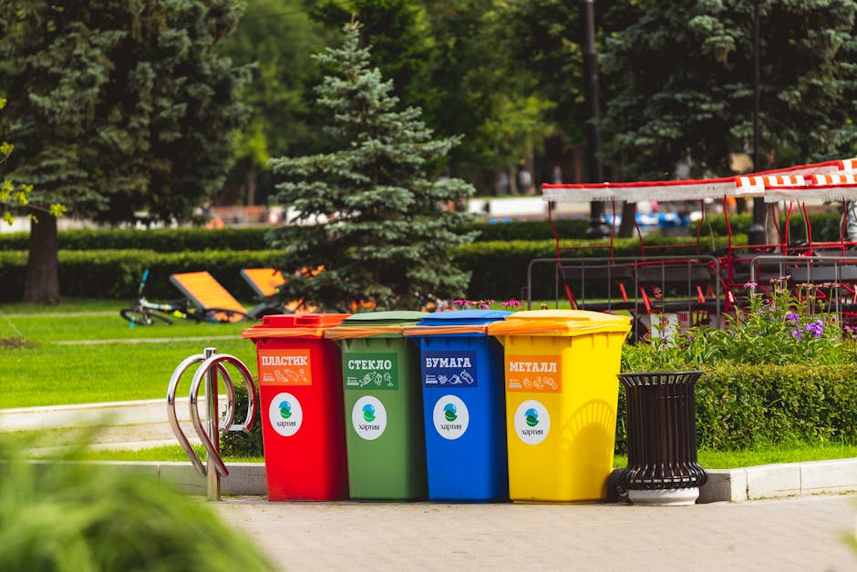 A street scene featuring a bright orange waste collection truck labeled with Vietnamese text, parked next to a sidewalk with a large tree in front. Several sanitation workers, dressed in yellow reflec