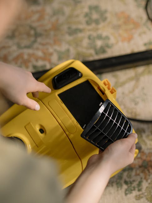 A person operating a yellow portable carpet cleaning machine, holding the device steady with both hands. The machine features a black vent or filter at the top front and a screen on the handle, indicating a modern carpet cleaning tool. The background includes a patterned carpet with muted colours and a portion of a light-colored wooden floor, reflecting cleanliness and maintenance. The lighting appears natural, highlighting the surface being cleaned, with no visible dust or stains, suggesting a thorough deep cleaning process. This image demonstrates surface cleaning and deep carpet maintenance, which is part of professional domestic or commercial sanitisation services offered by Cleaners Cleaning NW5 in the Kentish Town Road NW5 area.