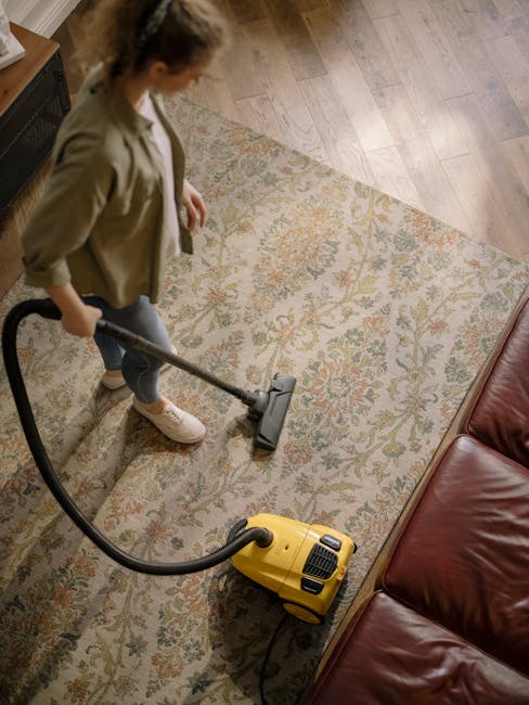 A woman performing surface cleaning in a living room, vacuuming a traditional patterned rug with a yellow vacuum cleaner. She is wearing casual clothing, including a beige jacket and jeans, and stands on a wooden floor adjacent to the rug. The room features a leather sofa positioned along the right side and ambient lighting that highlights the cleanliness and freshness of the space. The scene reflects a routine deep cleaning process, demonstrating the use of household tools to maintain hygiene, with the cleaning conducted by Cleaners Cleaning NW5, a specialist in domestic cleaning services.