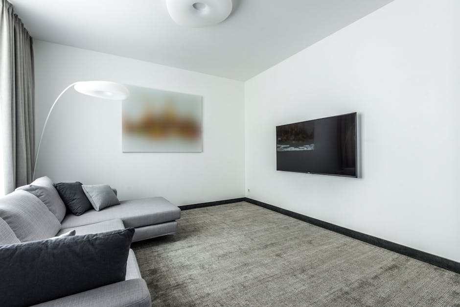 A modern living room with white walls and a grey carpeted floor, featuring a grey fabric sofa with dark and light cushions positioned along the left wall. An arched floor lamp with a white lampshade is situated next to the sofa, casting soft light across the space. On the wall behind the sofa, a blurred abstract artwork hangs, adding subtle color contrast. Opposite the sofa, a flat-screen television is mounted on the wall, reflecting ambient light. The room is illuminated by natural light from a window partially covered by a grey curtain on the left side. The overall cleanliness of the room suggests recent or ongoing surface cleaning and maintenance activities, consistent with services offered by Cleaners Cleaning NW5, emphasizing hygiene and deep cleaning standards for domestic surfaces.
