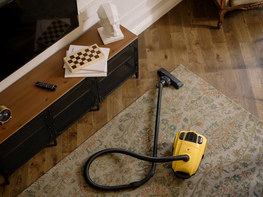 A woman performing surface cleaning in a living room, vacuuming a traditional patterned rug with a yellow vacuum cleaner. She is wearing casual clothing, including a beige jacket and jeans, and stands on a wooden floor adjacent to the rug. The room features a leather sofa positioned along the right side and ambient lighting that highlights the cleanliness and freshness of the space. The scene reflects a routine deep cleaning process, demonstrating the use of household tools to maintain hygiene, with the cleaning conducted by Cleaners Cleaning NW5, a specialist in domestic cleaning services.