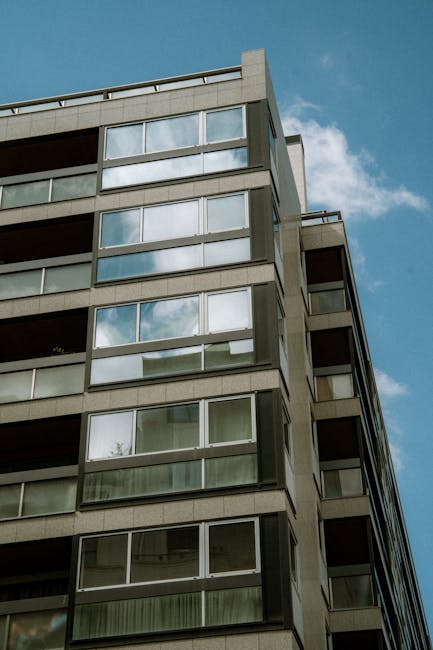 Photograph of a modern multi-storey residential building with a concrete façade and large glass windows, reflecting the blue sky and some clouds. The building features several balconies with metal railings, and the façade appears clean and well-maintained. Natural daylight illuminates the exterior, highlighting its structural details. This image illustrates the importance of surface cleaning and maintenance of external surfaces to preserve aesthetic appeal, relevant to residential cleans in the Crowndale Estate Kentish Town area, NW5, as provided by Cleaners Cleaning NW5.