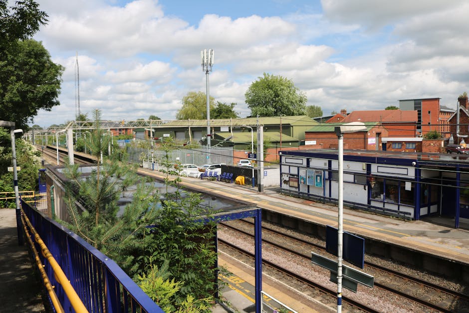 A view of Kentish Town station NW5 showing multiple railway tracks bordered by a blue safety railing on the platform side. The platform features a white and blue shelter with notices and public information boards attached. Surrounding the station are brick and green metal buildings, with trees and parked cars visible in the background. Overhead, electrical wires run parallel to the tracks, and a tall telecom pole stands near the station. The sky above is partly cloudy with patches of sunlight illuminating the scene, highlighting the clean, well-maintained surfaces of the station infrastructure as part of a professional cleaning and maintenance environment, in line with the services offered by Cleaners Cleaning NW5 for comprehensive surface and deep cleaning.