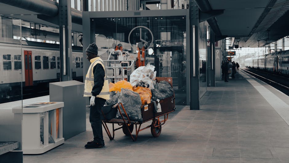 A worker at Kentish Town station NW5, part of the cleaning service by Cleaners Cleaning NW5, stands on the platform near a trolley loaded with plastic bags and cleaning equipment, including gloves, amid a spacious, well-lit train station. The station features modern metallic and glass architectural elements, with clean tiled flooring, stainless steel fixtures, and a waiting area in the background. The scene reflects a professional cleaning operation focusing on surface cleaning and sanitation, with an emphasis on hygiene and maintenance in a public transport environment.