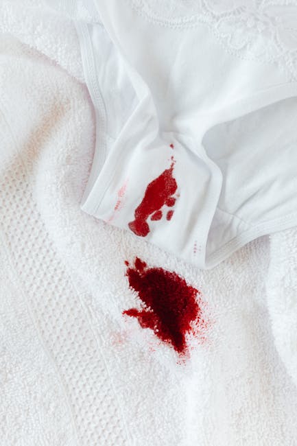 Close-up image of a white fabric surface, possibly a bedsheet or towel, featuring a noticeable red stain resembling a spilled liquid. The fabric appears soft with a textured pattern, and the stain is 