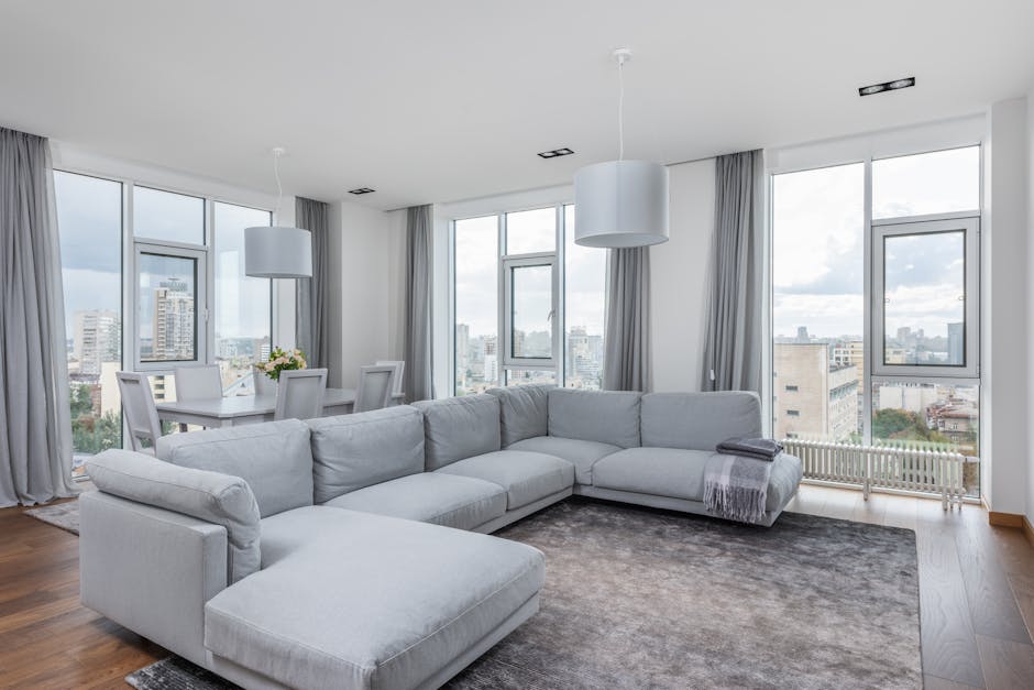 A spacious, modern living room with large floor-to-ceiling windows offering a view of the city skyline in Kentish Town NW5. The room features a light gray L-shaped sofa positioned on a dark gray area 