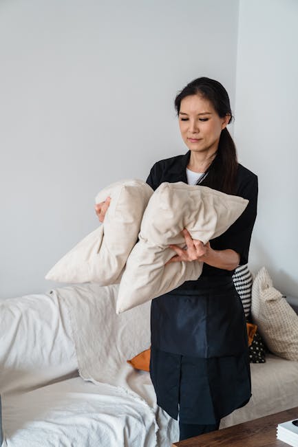 A professional cleaner from Cleaners Cleaning NW5 is preparing to perform surface cleaning and upholstery sanitisation in a residential living room within a domestic setting. The individual, dressed in a black uniform, is holding two beige cushions, indicating a focus on deep cleaning or refreshment of fabric surfaces. The room features a white sofa with a soft, clean appearance, and a side table with pillows, suggesting a comfortable, well-maintained space. Natural light illuminates the area, highlighting the cleanliness and orderliness of the surroundings. Visible cleaning tools include the cushions themselves, while no other cleaning equipment is present. The overall scene emphasizes expert domestic cleaning services aimed at maintaining hygiene and freshness in a home environment. For professional upholstery cleaning, contact Cleaners Cleaning NW5 for same-day service in the Kentish Town NW5 area, as shown on the page titled 'Same day upholstery cleaning Fortess Road Kentish Town, NW5.'</alt>